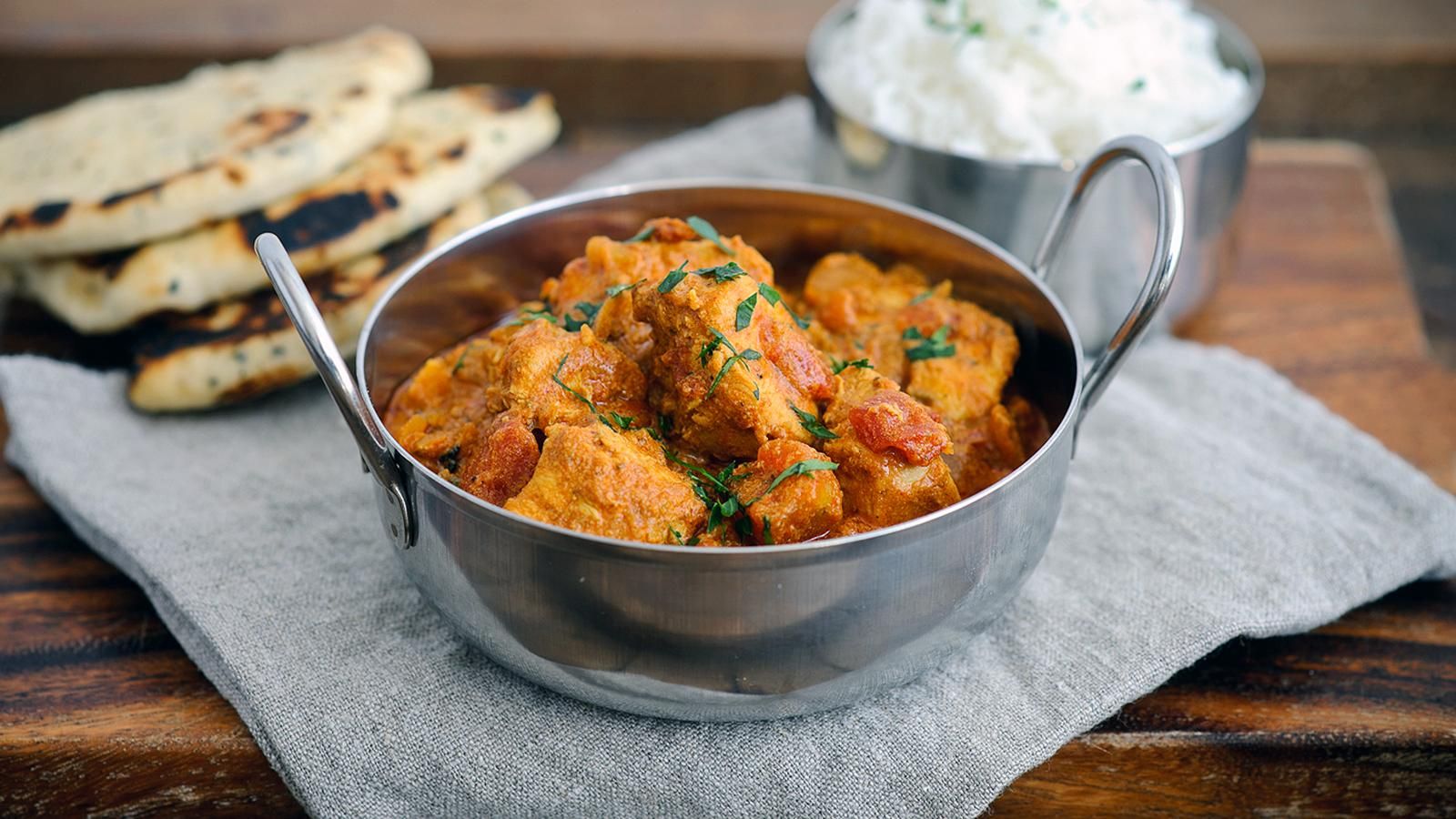 A bowl of homemade chicken tikka masala garnished with coriander, served with rice and naan bread on a rustic table.