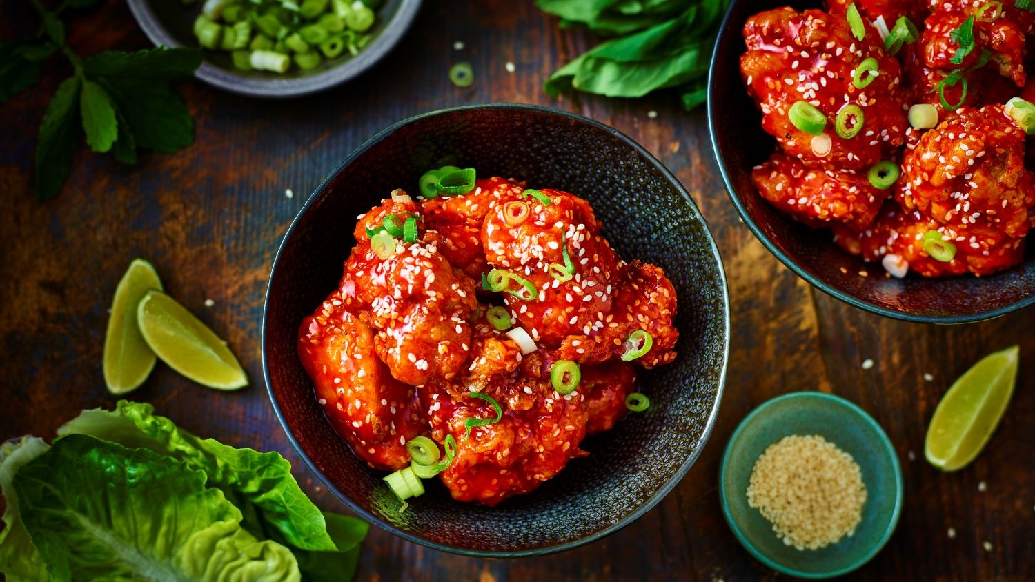 Two dark bowls filled with crispy Korean fried chicken coated in a glossy, spicy red sauce, garnished with white sesame seeds and chopped green onions, set on a rustic wooden table alongside fresh lime wedges and lettuce.