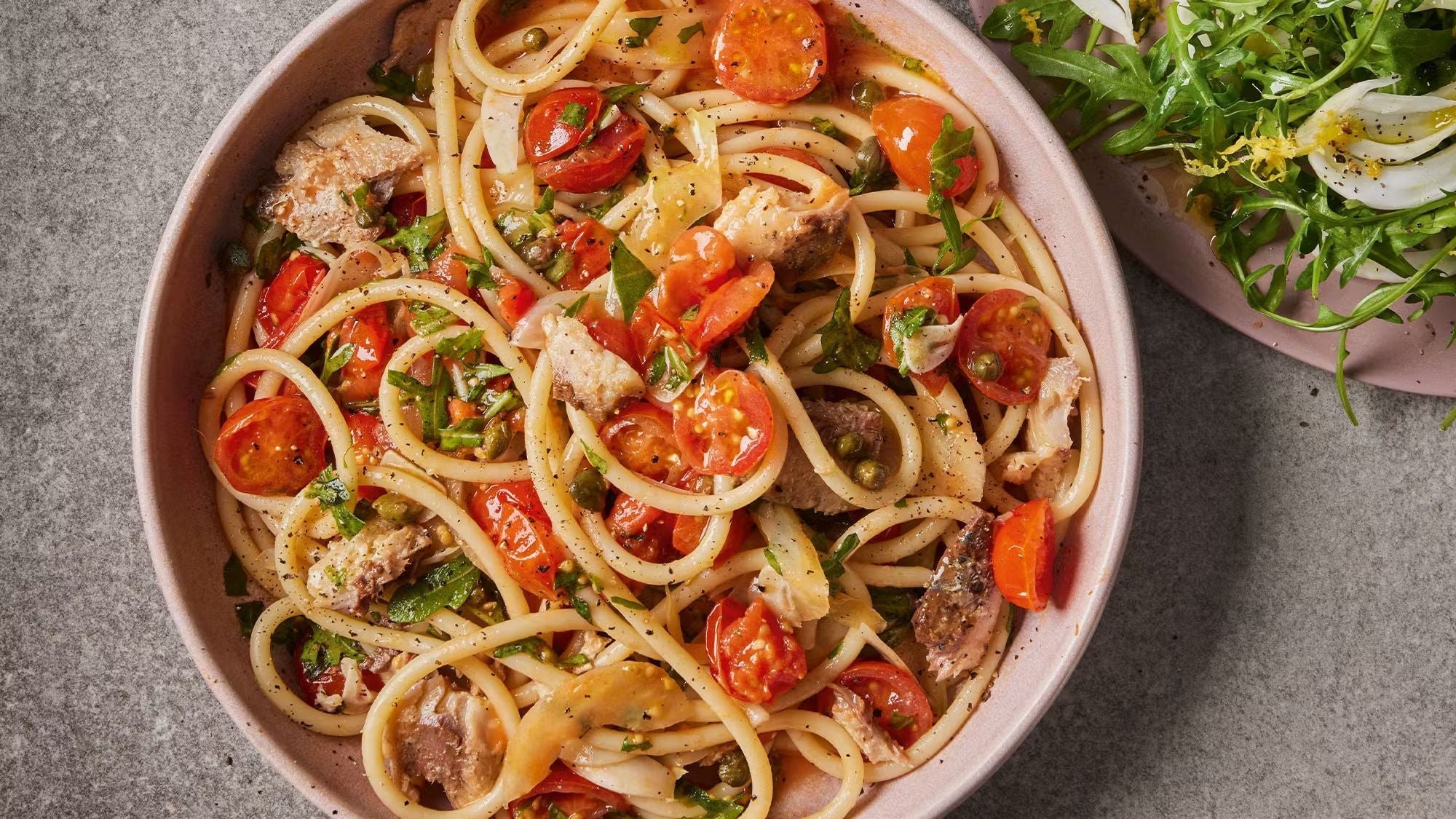 Bucatini pasta with sardines, cherry tomatoes, capers, and fresh herbs on a stone surface