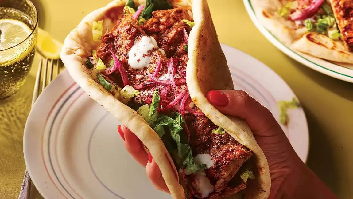 Hand holding spiced chicken kofta flatbread with yogurt, lettuce, and red onion