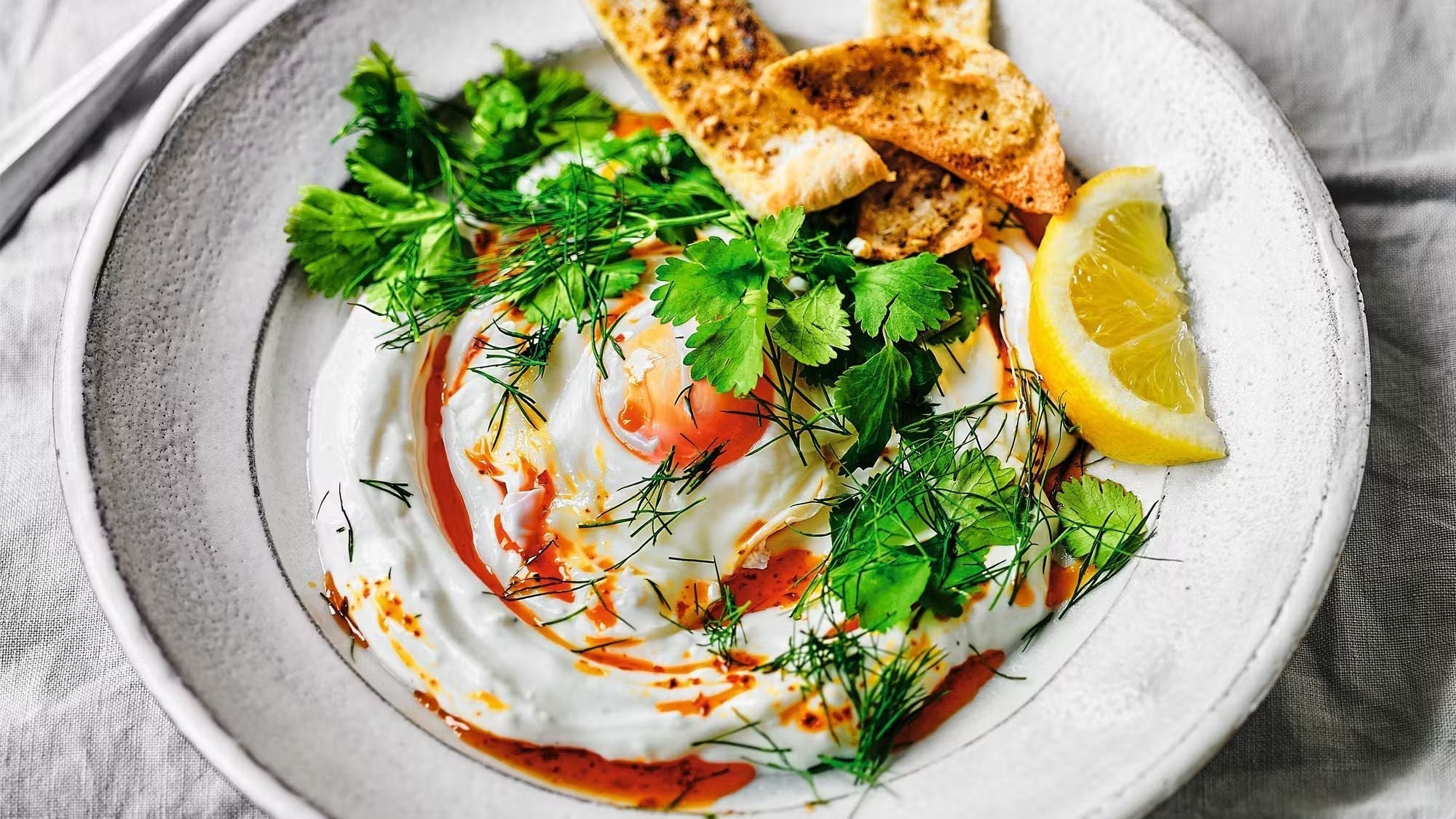 A close-up of Turkish Eggs (Çılbır) featuring a poached egg over creamy garlic yogurt, drizzled with vibrant pul biber oil, garnished with fresh dill and coriander, and served with toasted za'atar pitta crisps and a lemon wedge.
