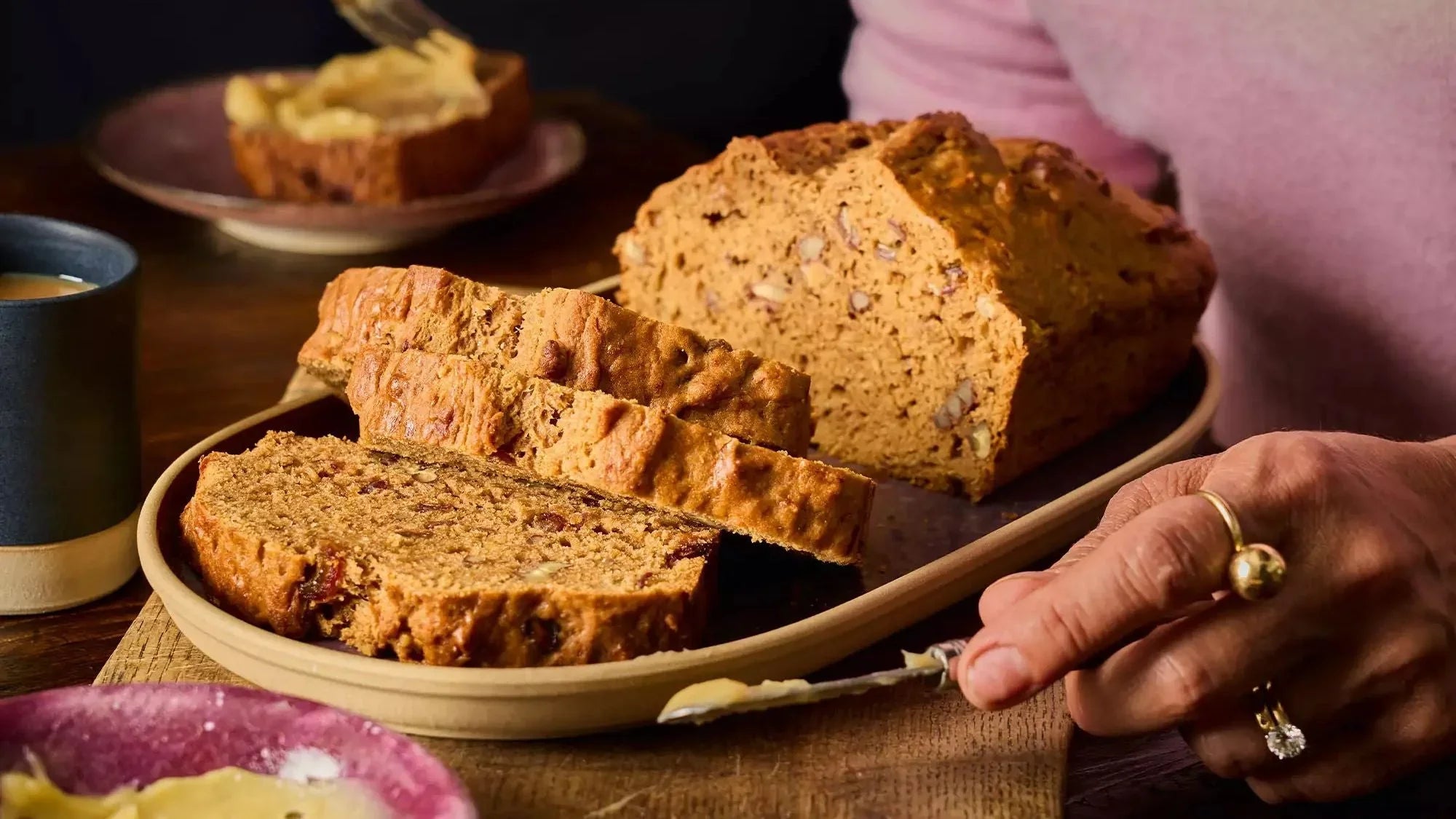 Sliced banana cake with visible nuts, served on a tray with honey butter and a cup of tea