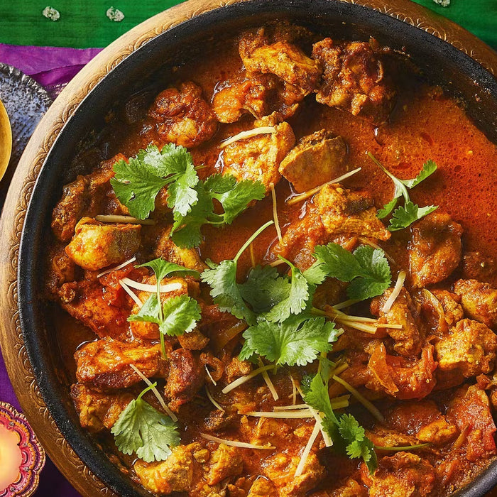 Traditional chicken karahi curry garnished with cilantro and ginger, served in a black pot with festive candles and sliced ginger on the side.
