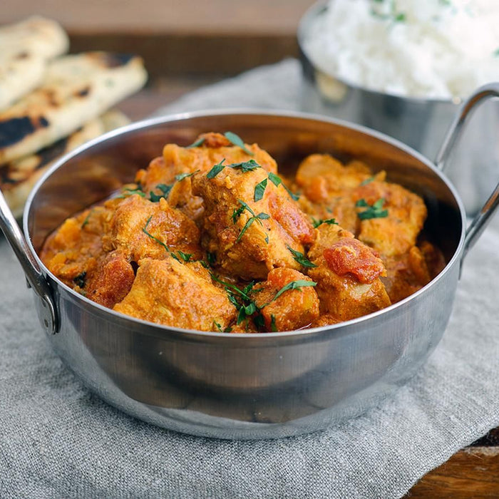 A bowl of homemade chicken tikka masala garnished with coriander, served with rice and naan bread on a rustic table.