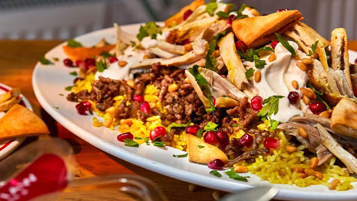 Fattet Dajaj dish with shredded chicken, yellow rice, pomegranate seeds, pine nuts, and fresh herbs on a white platter