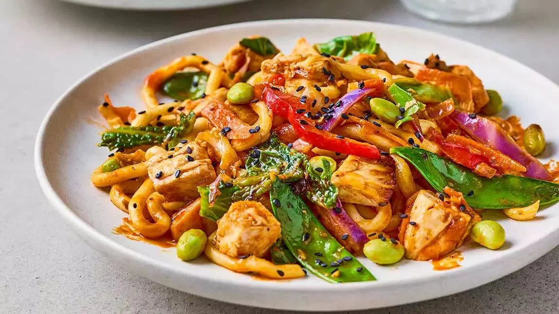 Gochujang butter chicken noodles with snap peas, red peppers, edamame, and black sesame on a white plate
