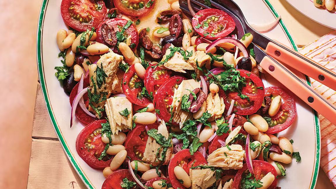 A vibrant summer salad on a white platter featuring ripe tomatoes, flaked tuna, creamy white cannellini beans, dark Kalamata olives, and fresh green herbs.