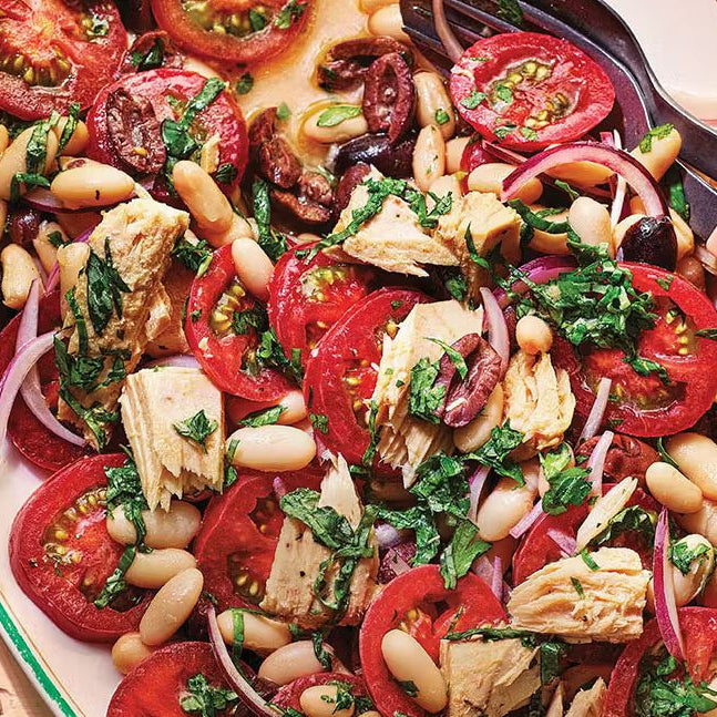 A vibrant summer salad on a white platter featuring ripe tomatoes, flaked tuna, creamy white cannellini beans, dark Kalamata olives, and fresh green herbs.