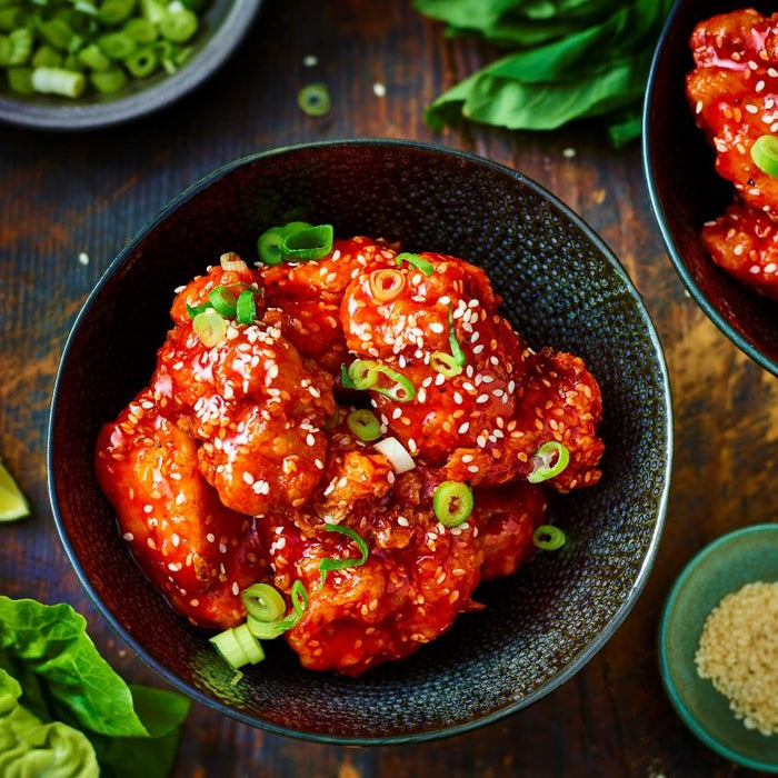 Two dark bowls filled with crispy Korean fried chicken coated in a glossy, spicy red sauce, garnished with white sesame seeds and chopped green onions, set on a rustic wooden table alongside fresh lime wedges and lettuce.