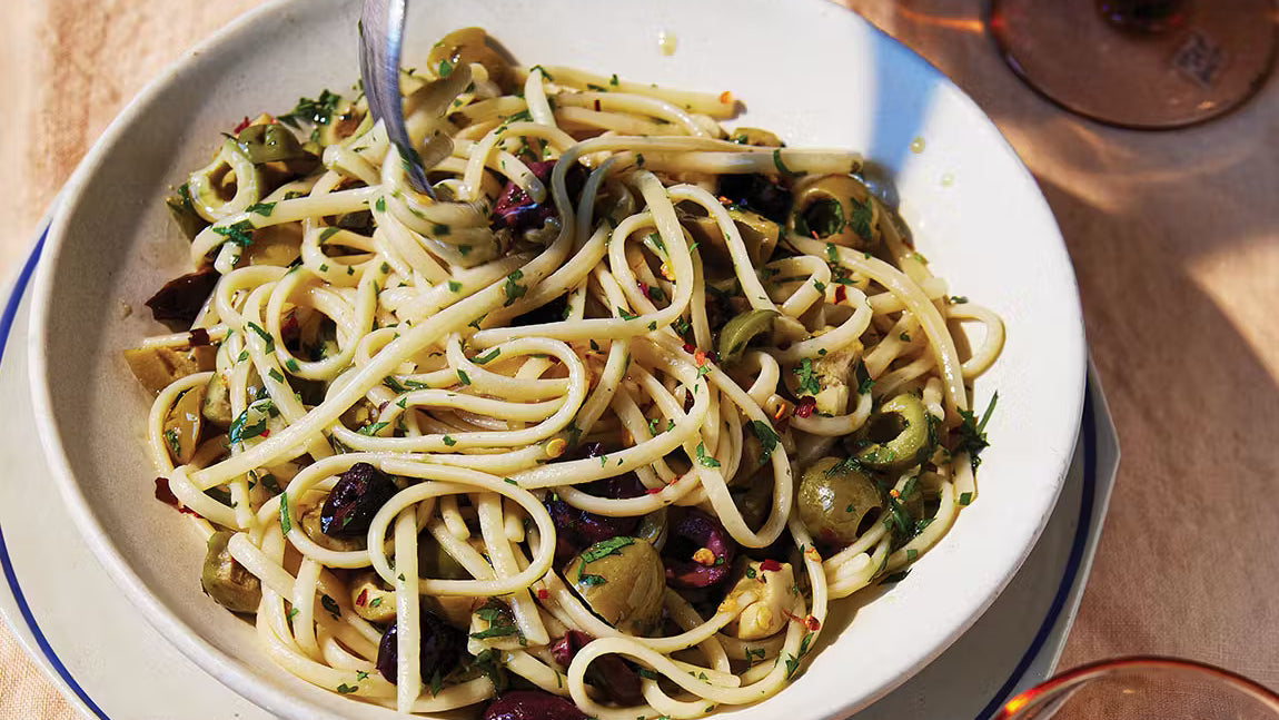 Linguine aglio e olio pasta with green and black olives, herbs, and chili flakes in a white bowl.