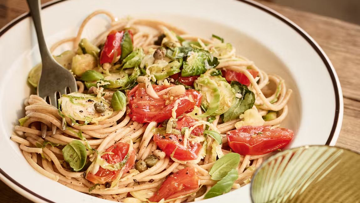 A bowl of wholewheat spaghetti tossed with roasted plum tomatoes, crispy shredded Brussels sprouts, capers, and fresh basil leaves.