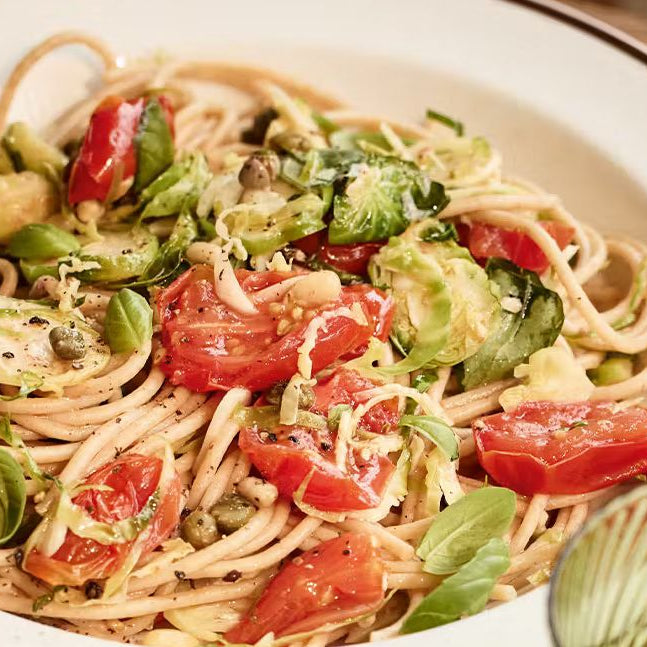 A bowl of wholewheat spaghetti tossed with roasted plum tomatoes, crispy shredded Brussels sprouts, capers, and fresh basil leaves.