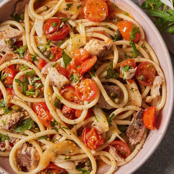 Bucatini pasta with sardines, cherry tomatoes, capers, and fresh herbs on a stone surface