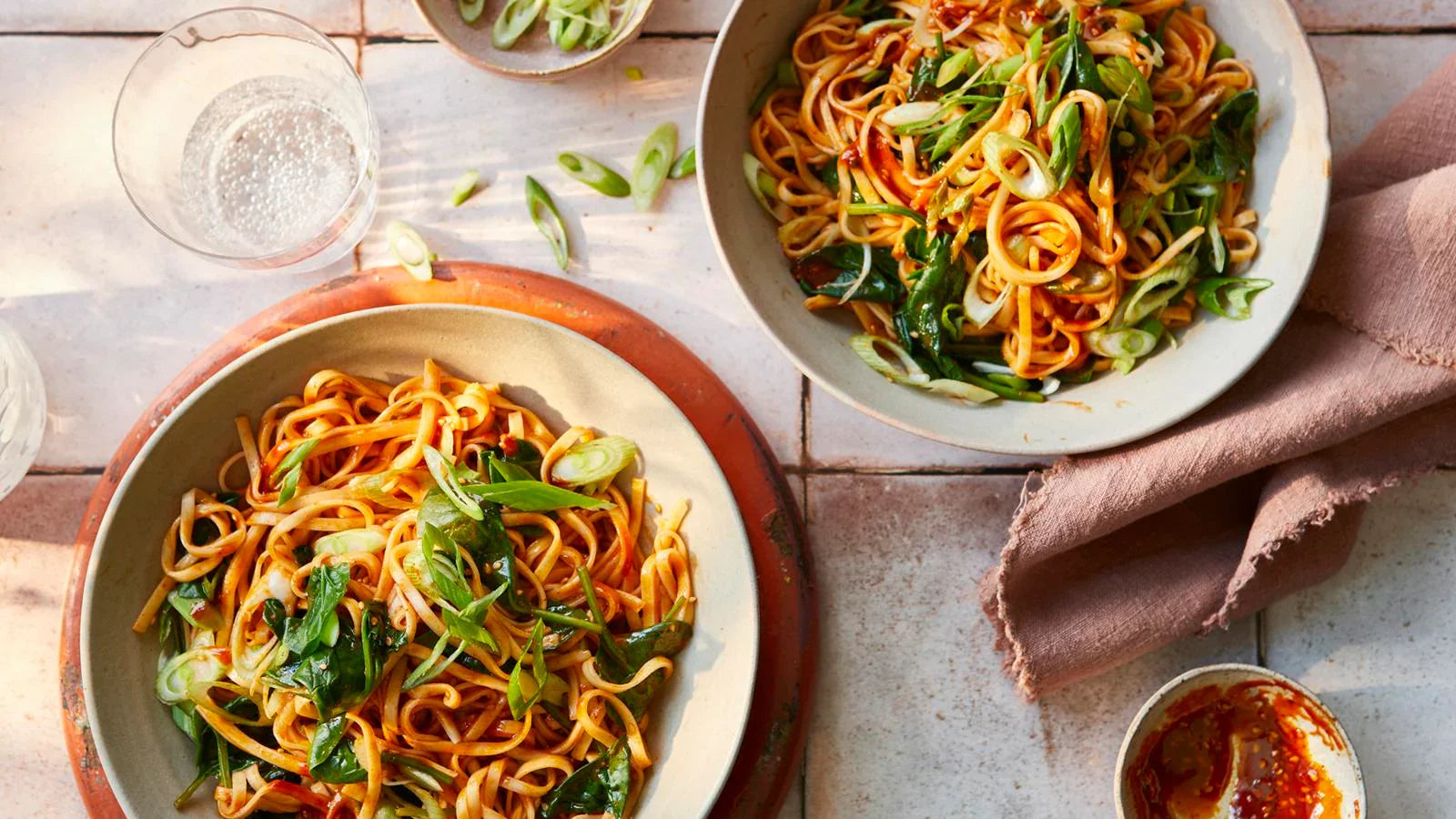 Bowls of spicy gochujang noodles topped with fresh greens and spring onions on a rustic table.