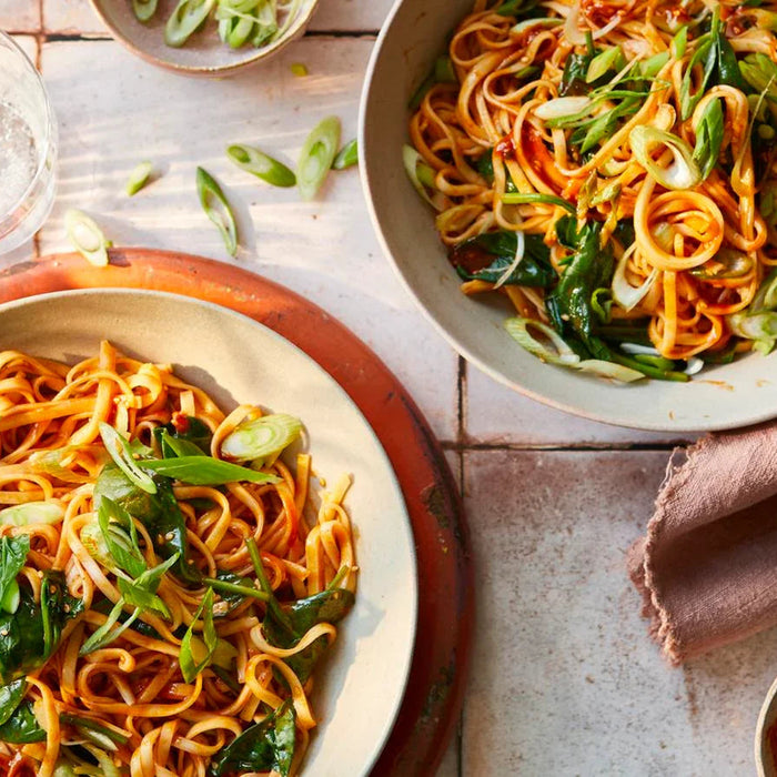 Bowls of spicy gochujang noodles topped with fresh greens and spring onions on a rustic table.