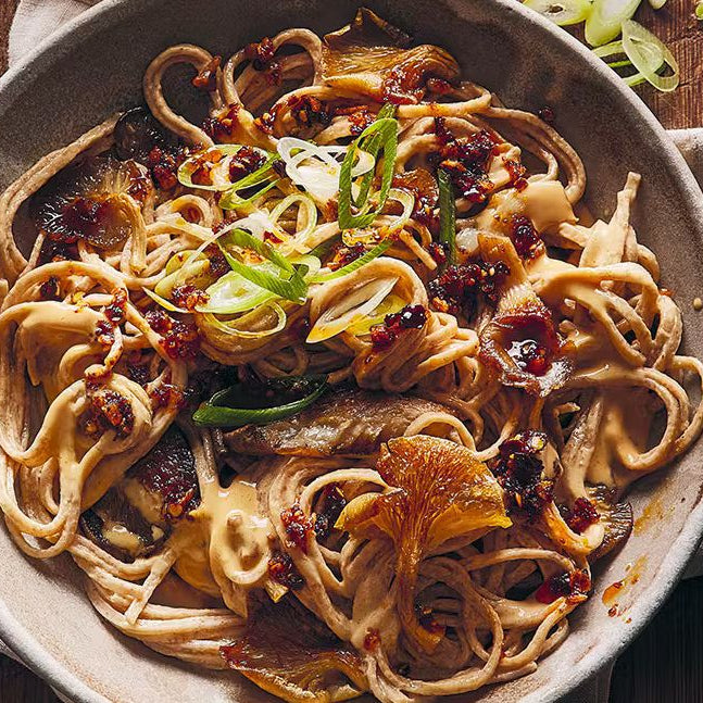 Bowl of sesame noodles topped with sticky mushrooms, chili oil, and sliced green onions