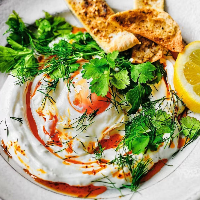 A close-up of Turkish Eggs (Çılbır) featuring a poached egg over creamy garlic yogurt, drizzled with vibrant pul biber oil, garnished with fresh dill and coriander, and served with toasted za'atar pitta crisps and a lemon wedge.
